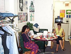 Pic: A messy table with Deb and Spring reading. There are posters on the walls, clothes hanging on a rack. On the table is food and bottled water.