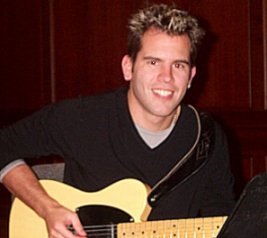 A handsome Shawn Cooney holding his guitar. He has a fresh all-American face and a great big smile.